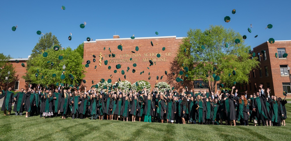 Large group of people in college gowns standing in front of a WVSOM sign and throwing their graduation hats into the air.