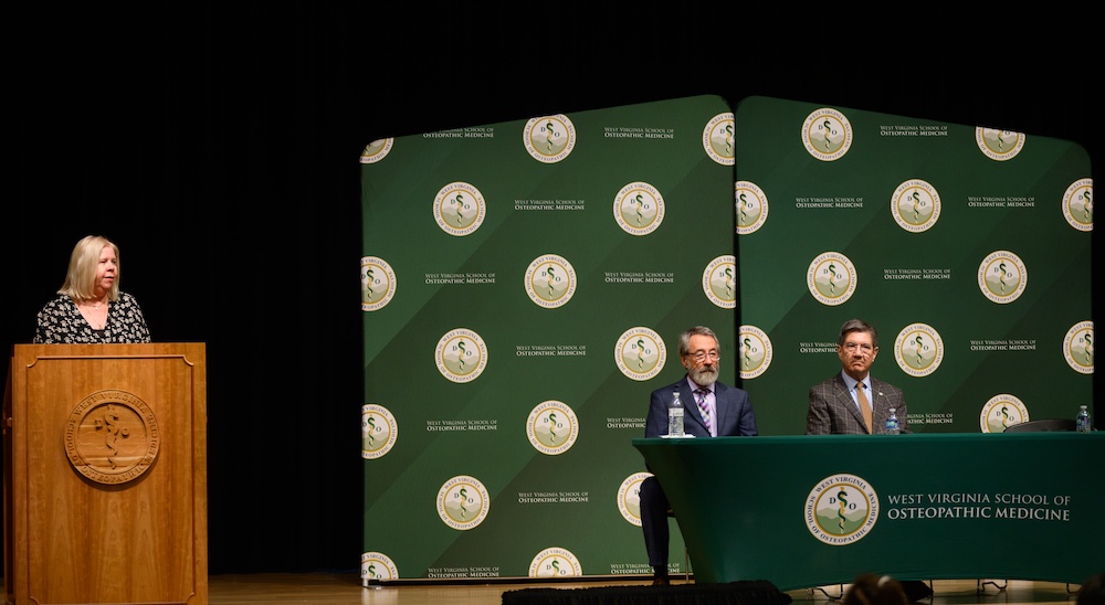 Woman talks at a podium while two men sit and listen.