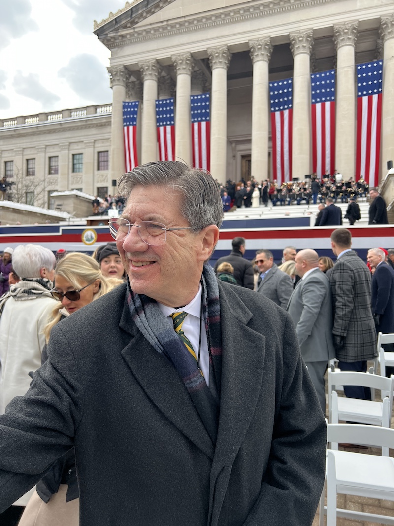 Man in coat stands outside of capitol building with flag displays