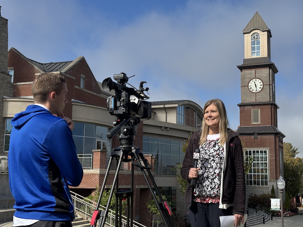 woman speaks to a news camera while a man records her.