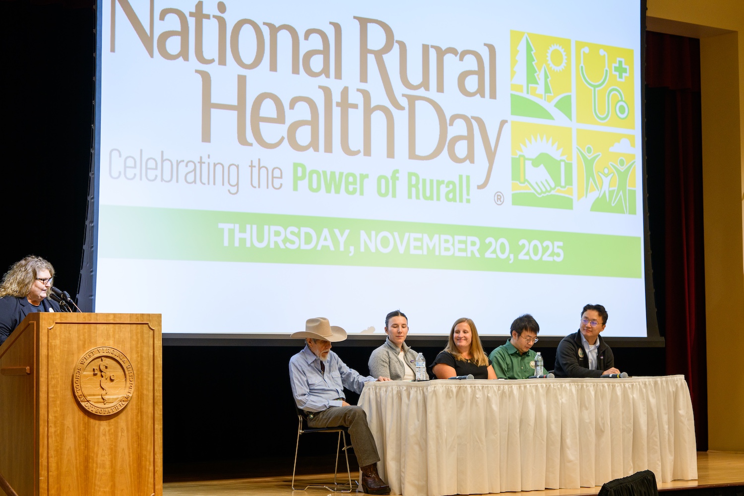 Woman at podium and five people seated in front a screen.