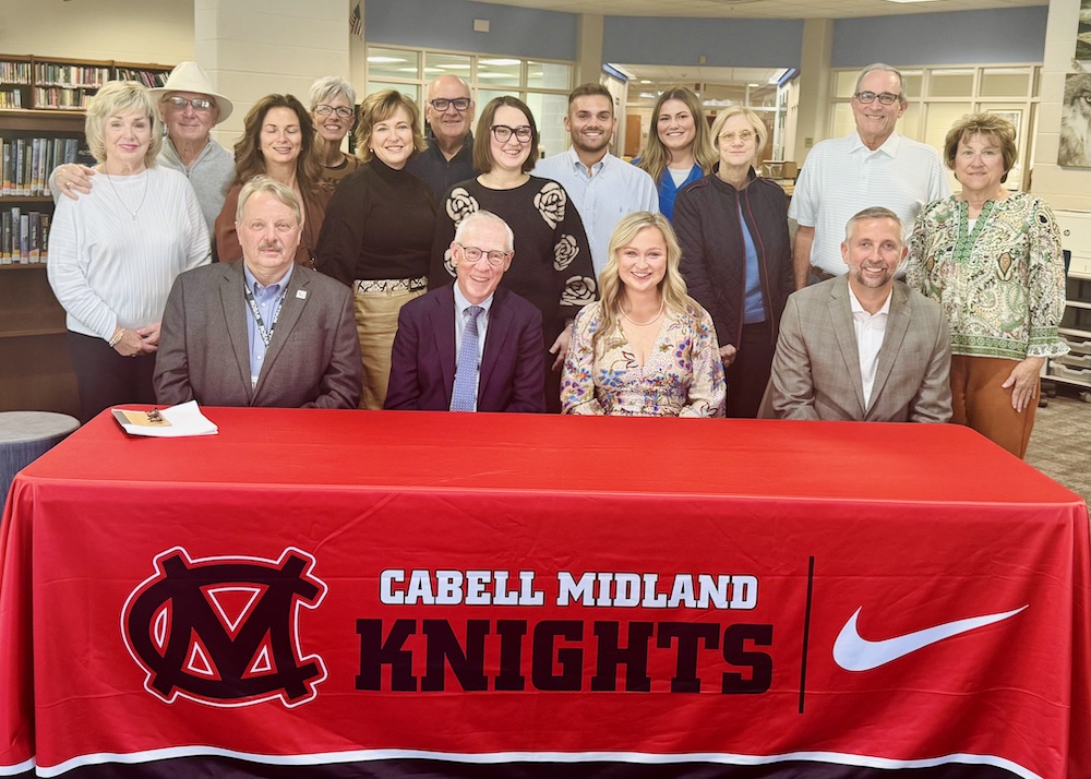 Four people sit at a table and 12 stand behind them. 