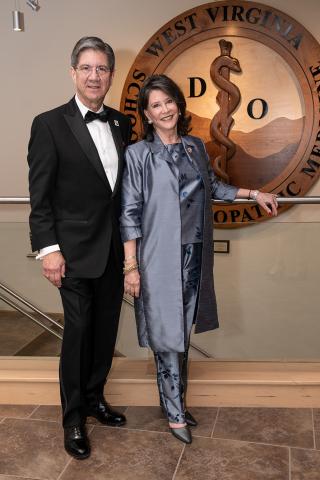 Dr. Nemitz and wife Nancy, dressed in formal attire,  pose in front of wooden WVSOM logo