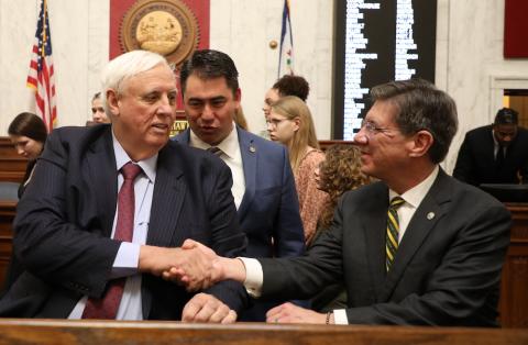 Two men shake hands and a third man watches the exchange in a crowded room 