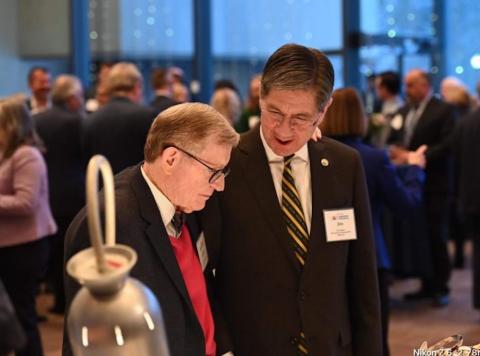 two men lean in to talk in a crowded room