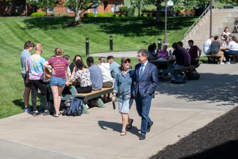 Woman and a man walking outdoors near people seated at a table.