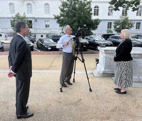 Woman stands in front of camera and camera man while another man watches. 