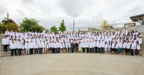 Two hundred students wearing white coats stand outside on steps. 