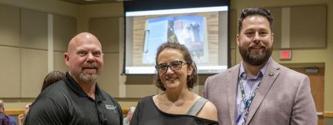 A man, woman and man stand in a line with a screen behind them. 