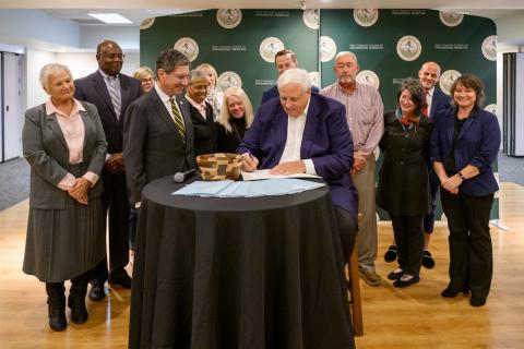 Group of people stand around one man signing a document.