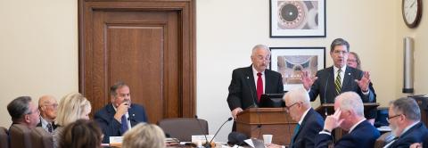 Two men stand. One talks to people in a meeting.