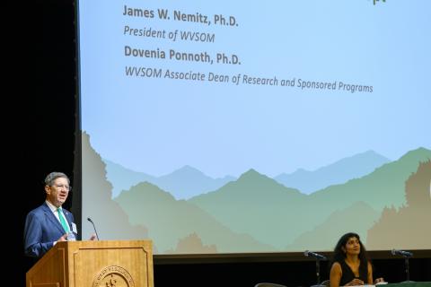 Man stands at podium and woman sits at table. 