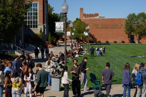 A large group of people stand along a building and grassy lawn on a sunny day. 