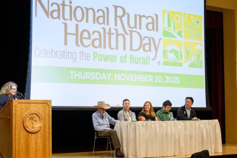 Woman at podium and five people seated in front a screen.