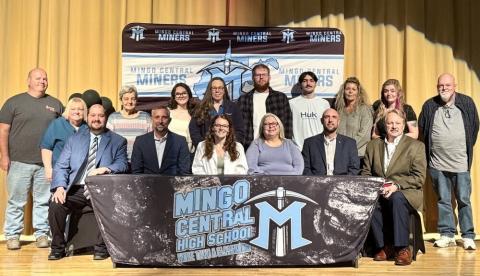 six people sit at a table in front of 10 people standing on a stage.