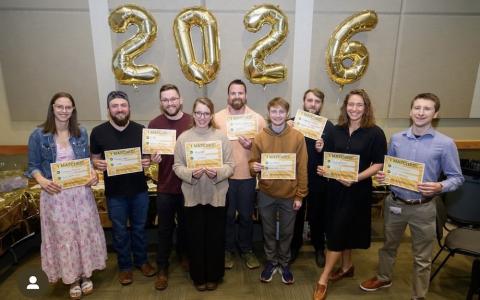 Nine men and women hold signs and stand in front of "2026" balloon