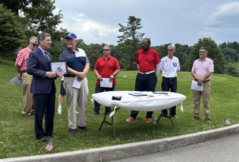 Man stands with an award and speaks while other men look observe his comments. 