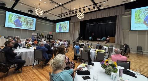 Large conference with people seated at tables watching video screens.
